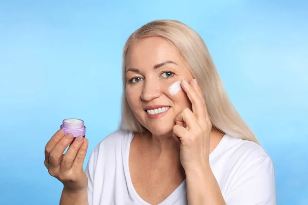 Mujer madura aplicando crema facial sobre fondo de color — Foto de Stock