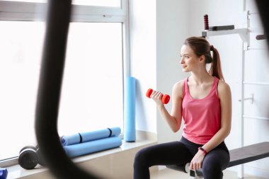 Spor salonunda dambıllarla spor yapan genç bir bayan.