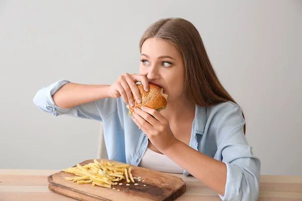 Young woman eating tasty burger with french fries at table — Stock Photo, Image