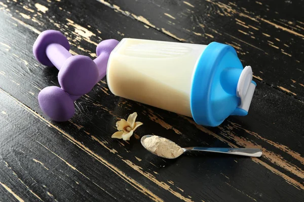 Bottle of protein shake with dumbbells on wooden table