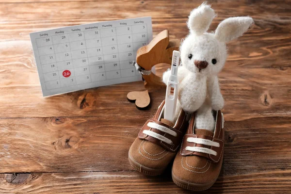 Baby booties, toy rabbit, pregnancy test and calendar on wooden background — Stock Photo, Image