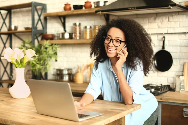 Female African-American freelancer talking by phone while working on laptop in kitchen — Stock Photo, Image