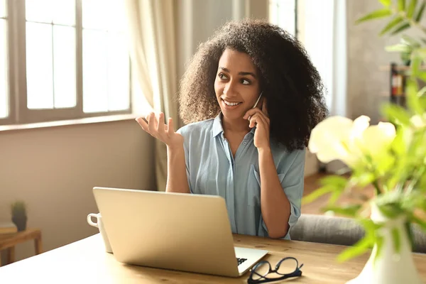 Female African-American freelancer talking by phone while working on laptop at home — Stock Photo, Image