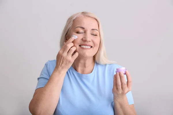 Mulher madura aplicando creme facial no fundo cinza — Fotografia de Stock