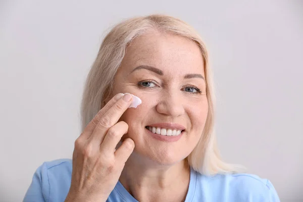 Mujer madura aplicando crema facial sobre fondo gris — Foto de Stock