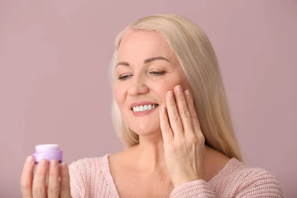 Mature woman applying facial cream on color background — Stock Photo, Image
