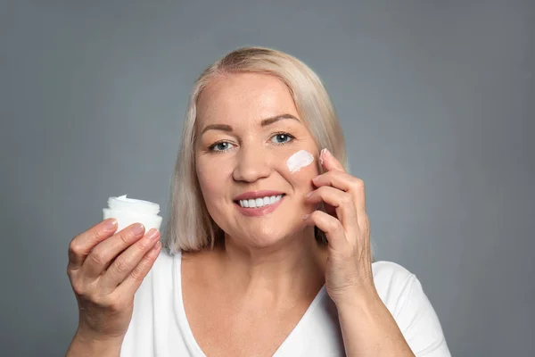 Mujer madura aplicando crema facial sobre fondo gris — Foto de Stock