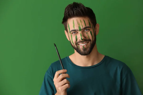 Portrait of handsome man with paint on his face holding brush against color background — Stock Photo, Image