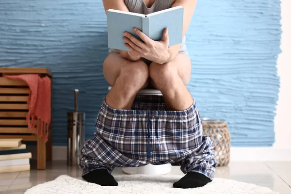 Man reading book while sitting on toilet bowl at home — Stock Photo, Image