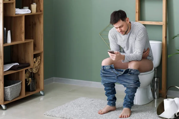 Homem com telefone celular sofrendo de diarreia enquanto sentado em vaso sanitário em casa — Fotografia de Stock