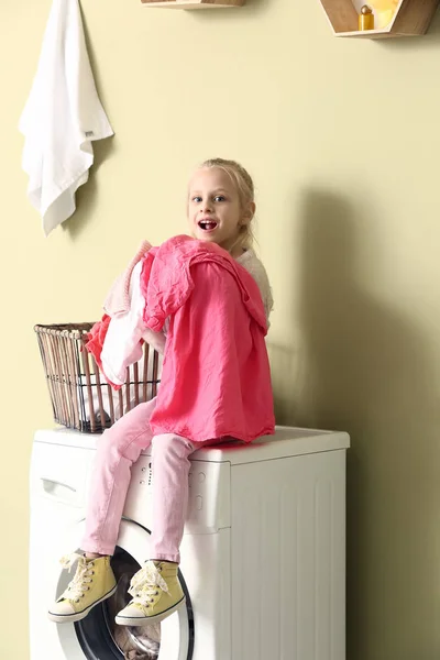 Little girl with laundry sitting on washing machine at home — Stock Photo, Image