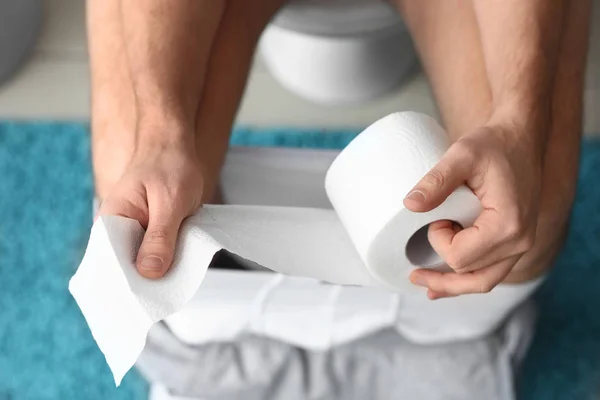 Man with roll of paper sitting on toilet bowl at home, closeup — Stock Photo, Image