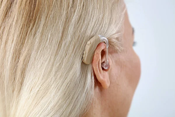 Mature woman with hearing aid on light background, closeup