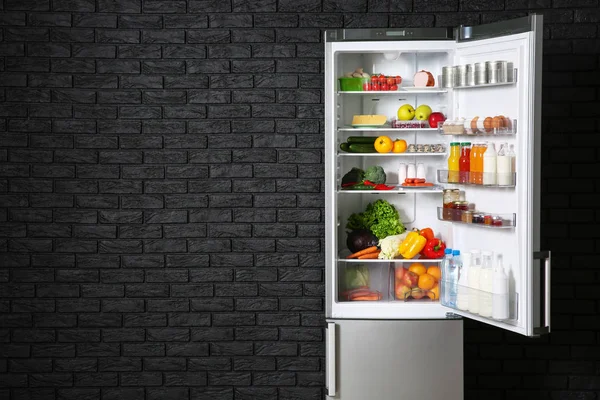 Відкритий холодильник повний їжі біля темної цегляної стіни — стокове фото