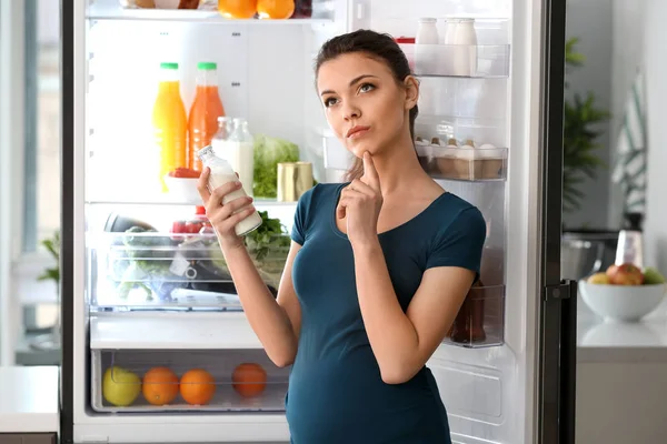 Задумчивая беременная женщина стоит у открытого холодильника дома — стоковое фото