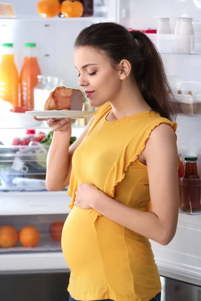 Беременная женщина с вкусным мясом стоит у открытого холодильника дома — стоковое фото