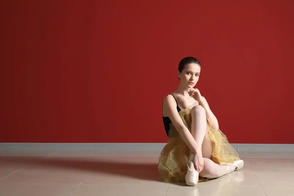 Beautiful young ballerina sitting on floor against color wall