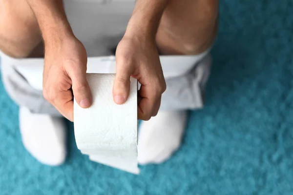 Man with roll of paper sitting on toilet bowl at home, closeup — Stock Photo, Image