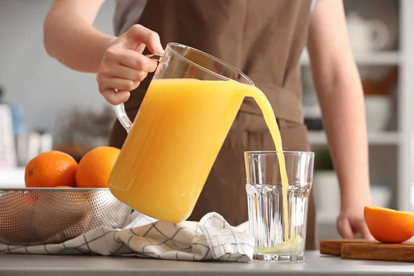 Woman pouring orange juice from jug into glass in kitchen — Stock Photo, Image