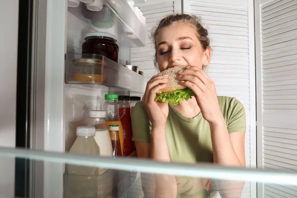 Mulher bonita comendo comida perto da geladeira, vista de dentro — Fotografia de Stock