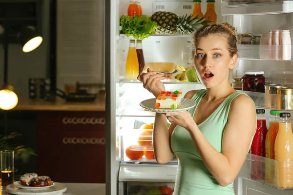 Mulher com medo pego no ato de comer comida insalubre saborosa perto da geladeira à noite — Fotografia de Stock