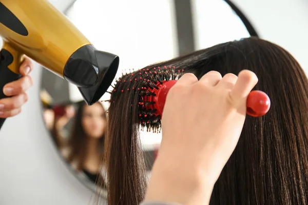 Hairdresser with blow dryer brushing long hair of young woman in salon — Stock Photo, Image