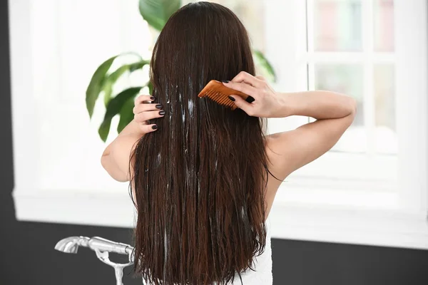 Mulher bonita aplicando cosméticos em seu cabelo longo saudável em casa — Fotografia de Stock