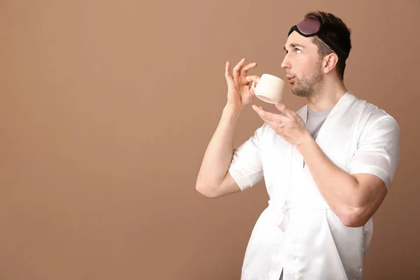 Funny young man with sleep mask and cup of coffee on color background