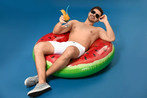 Handsome man with cocktail relaxing on inflatable ring against color background