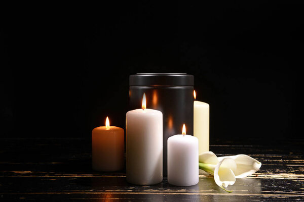 Mortuary urn, burning candles and flowers on table against dark background