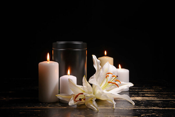 Mortuary urn, burning candles and flowers on table against dark background
