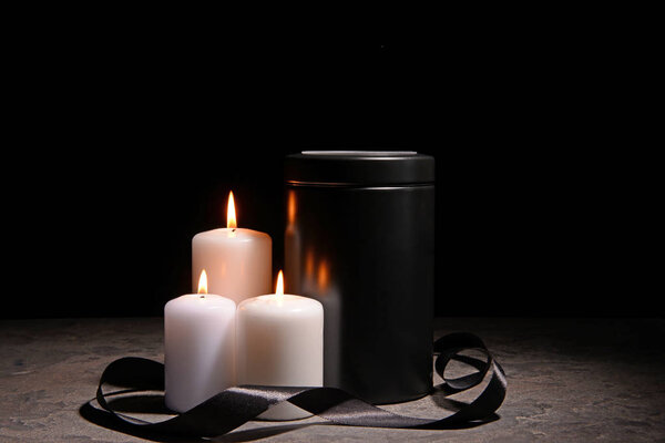 Mortuary urn, burning candles and black ribbon on table against dark background