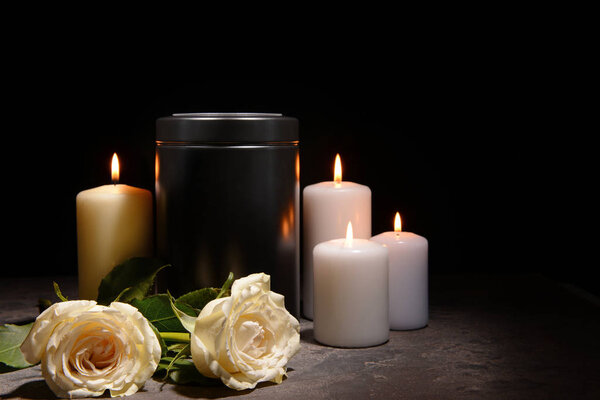 Mortuary urn, burning candles and flowers on table against dark background