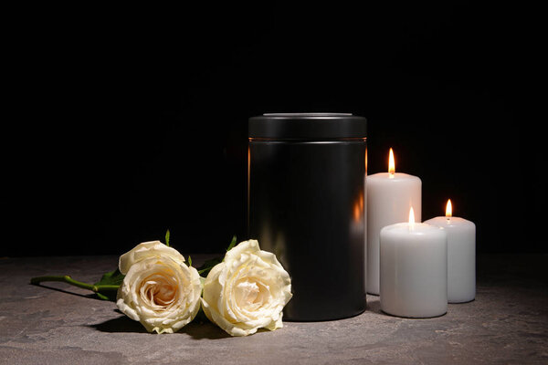 Mortuary urn, burning candles and flowers on table against dark background