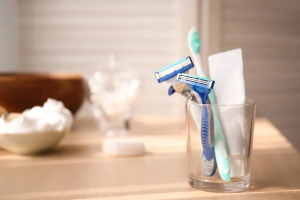 Glass with razors, tooth paste and brush on table in bathroom — Stock Photo, Image