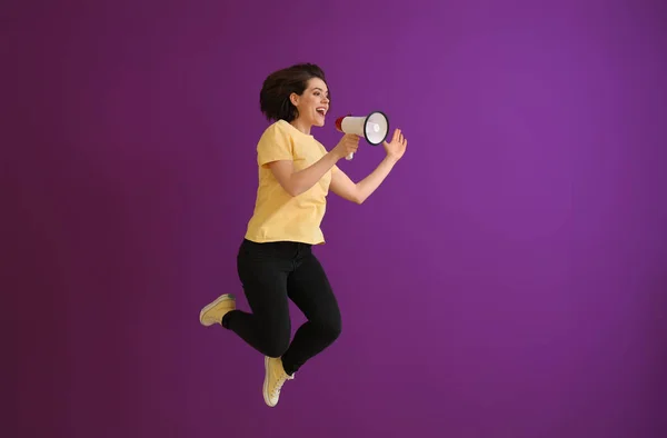 Jumping young woman with megaphone against color background — Stock Photo, Image