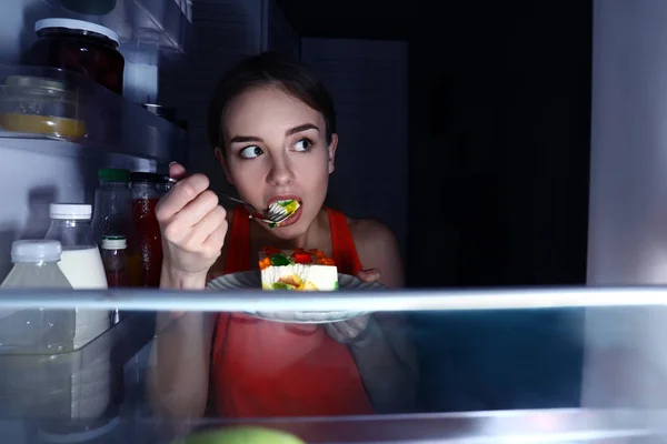 Mulher jovem desconfiada comendo comida perto da geladeira, vista de dentro — Fotografia de Stock