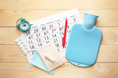 Ahşap arka planda kadınsı ürünler, haplar, su torbası ve çalar saat ile menstrüel takvim