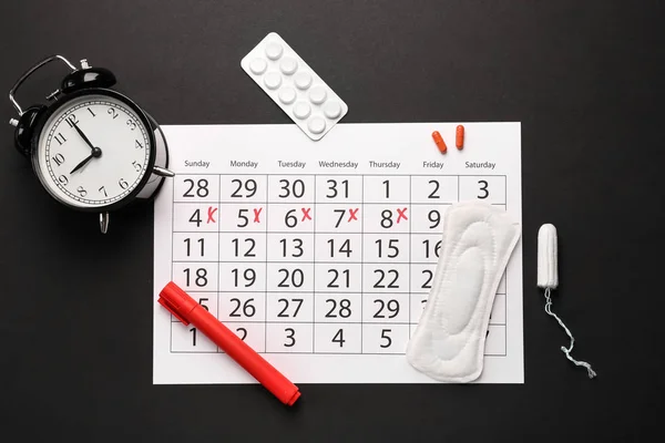 Menstrual calendar with feminine products, pills and alarm clock on dark background — Stock Photo, Image