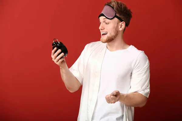 Happy young man with sleep mask and alarm clock on color background — Stock Photo, Image