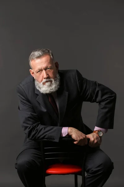 Handsome mature businessman sitting on chair against dark background — Stock Photo, Image