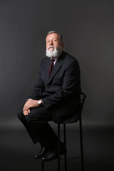Handsome mature businessman sitting on chair against dark background — Stock Photo, Image