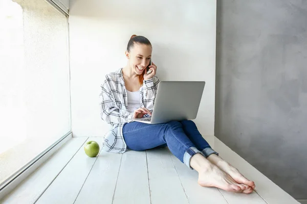 Young woman talking by phone while working on laptop indoors — Stock Photo, Image