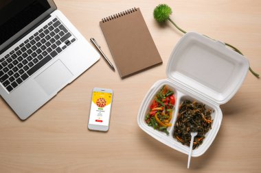 Ekranda gıda dağıtım sitesi açık sayfa ile Cep telefonu, ahşap masada dizüstü bilgisayar ve paket yemek