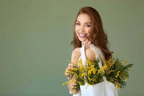 Beautiful young woman holding bag with mimosa flowers on color background