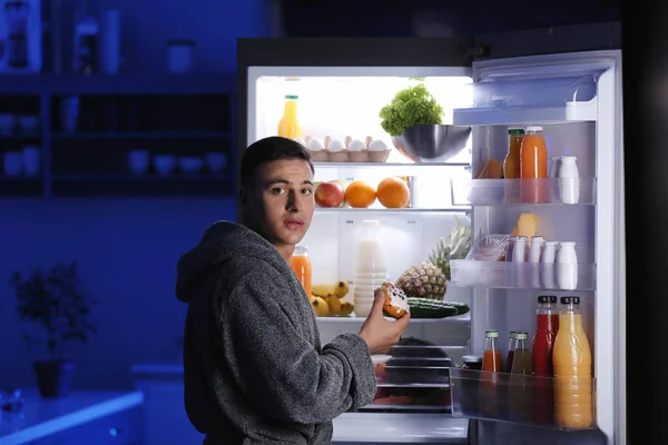 Afraid man caught in the act of eating unhealthy food near refrigerator at night