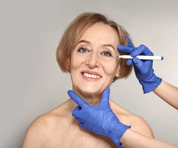 Surgeon drawing marks on face of mature woman against grey background — Stock Photo, Image