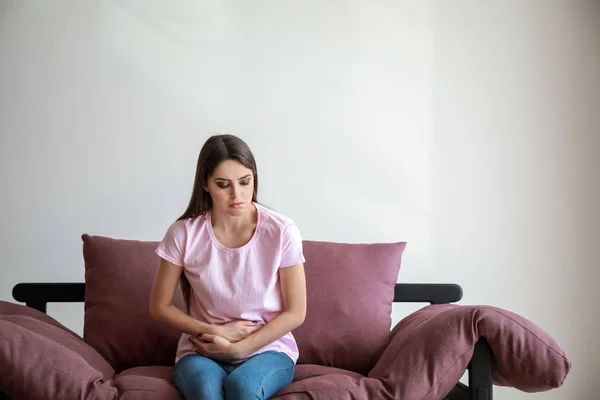 Fiatal nő szenved hasi fájdalom, miközben ül a kanapén szemben fehér háttér — Stock Fotó