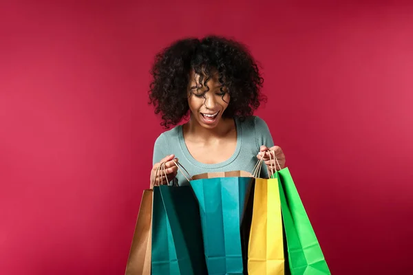 Happy African-American woman looking into shopping bag on color background — Stock Photo, Image