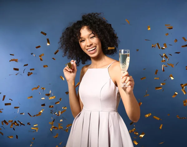 Happy African-American woman with glass of champagne and falling confetti on color background — Stock Photo, Image